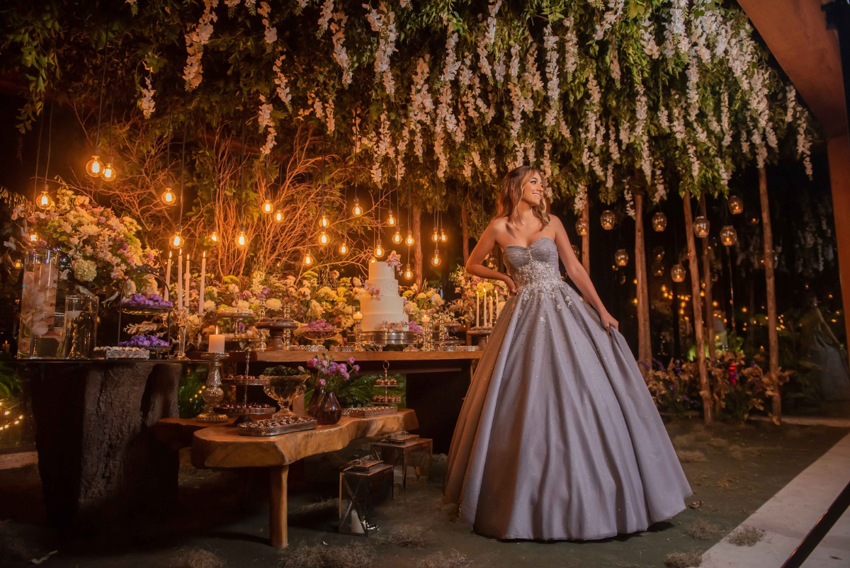A debutante posa ao lado da mesa de doces, decorada com arranjos florais, velas e luminárias suspensas em meio à vegetação | Foto: Bruno Felipe Fotografia